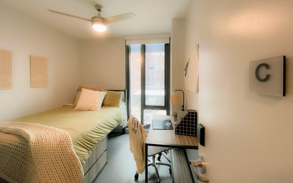 Student dorm room with green blankets and pillows on a bed, a desk and ceiling fan of a Georgetown apartment at 55 H Street