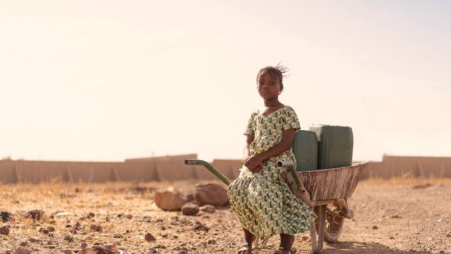 A child sits in a wheelbarrow surrounded by desert landscape.