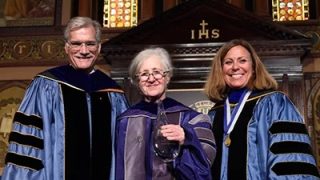 Maxine Weinstein, wearing academic robes, stands between Provost Robert Groves (left) and vice provost of research, Janet Mann (right) in 2015 to receive the Career Research Achievement Award in Gaston Hall