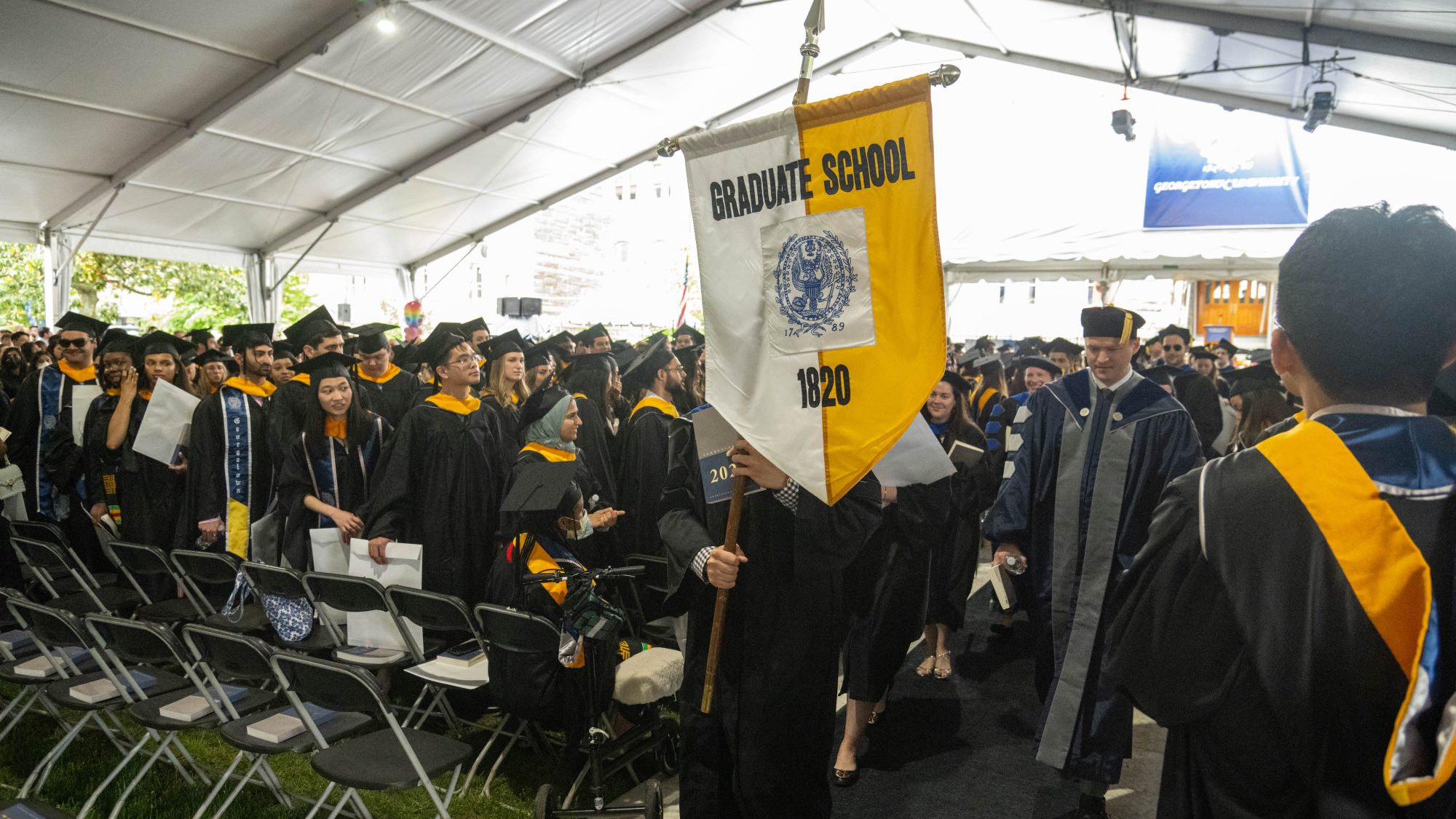 Graduate students wearing caps and gowns line Healy Lawn at commencement with the Graduate School banner (half white, L, and half yellow, R) being carried by a student during the 2022 recessional