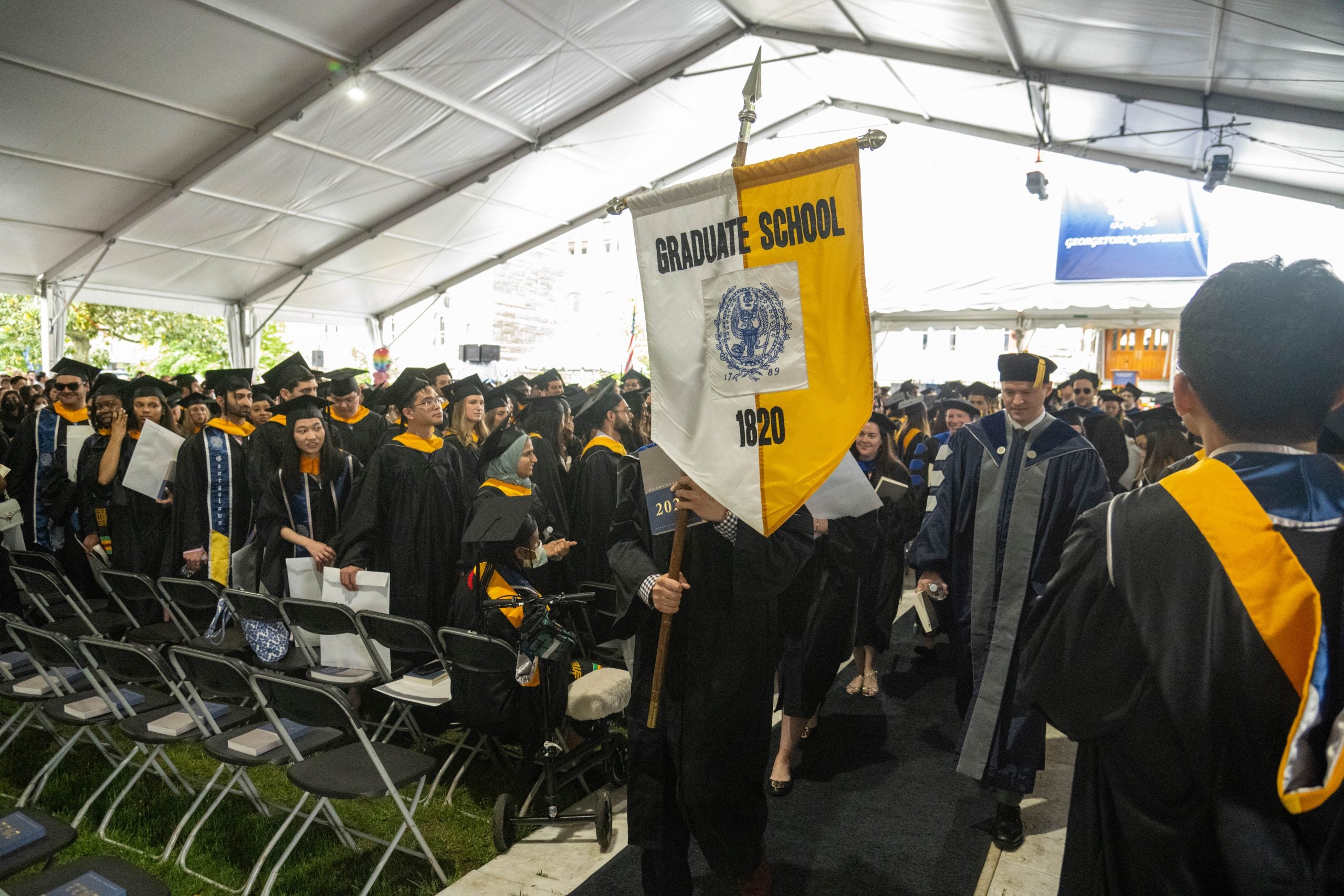 Graduate students wearing caps and gowns line Healy Lawn at commencement with the Graduate School banner (half white, L, and half yellow, R) being carried by a student during the 2022 recessional