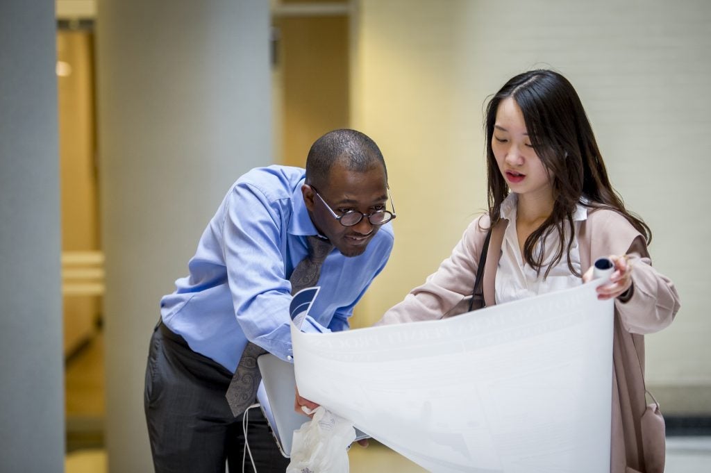 Two people standing close together (a Black male, left, and an Asian woman, right), peer over a drawing