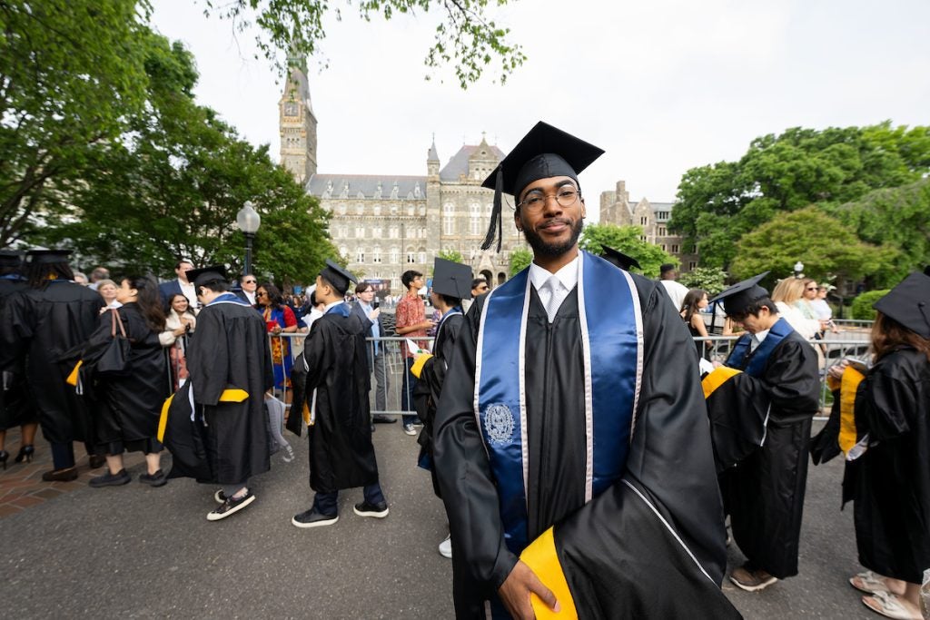 A student wearing graduation robes stands for his photo to be taken with Healy Hall and classmates processing into the 2023 Commencement Ceremony in the background