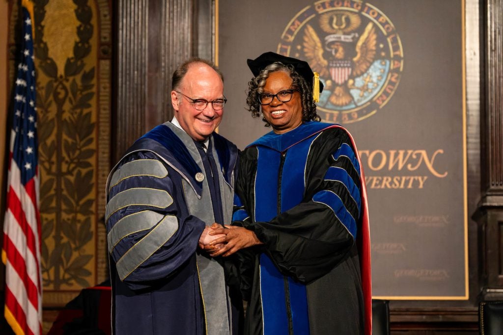 Dr. Lucile Adams-Campbell (right) shakes hands with Georgetown University President, Jack DeGioia (left); they are both wearing their academic robes and stand in front of the Georgetown seal and logo on the back wall inside Gaston Hall