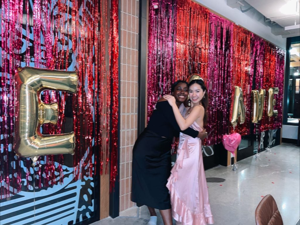 Two graduate students hug and pose for a photo at the Capital Campus Residence Hall 'Love Game' event in February 2023; behind them the walls are decorated in pink and red streamers with gold balloons