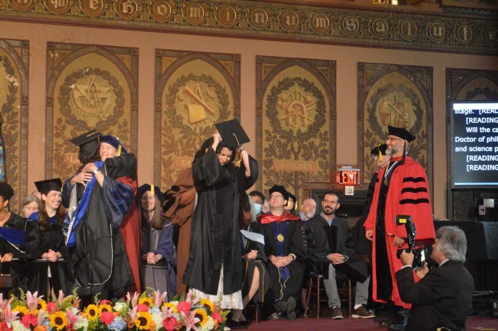 Two doctoral students have their hoods placed on them by faculty at the Doctoral Hooding Ceremony in 2023 on stage inside Gaston Hall; faculty members are seated behind them on stage and Dean Alexander Sens (right) stands and smiles at them dressed in his red academic regalia robes
