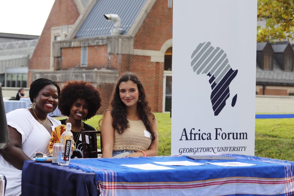 Three graduate student representatives of the Africa Forum (left) sit behind a table with a step-and-repeat banner (right) displaying the African continent with text that reads 'Africa Forum' at the 2023 Welcome Week BBQ on Leavey Esplanade