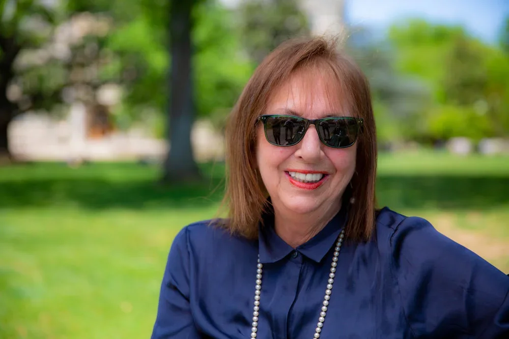 Headshot of Elizabeth Velez (G'83) wearing a blue blouse, pearls and sunglasses in front of a green background
