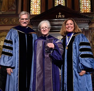 Georgetown University Provost Bob Groves (left), Professor Maxine Weinstein (center), and Professor Janet Mann (right) pose for a photo on stage inside Gaston Hall as Weinstein accepts an award, which she is holding