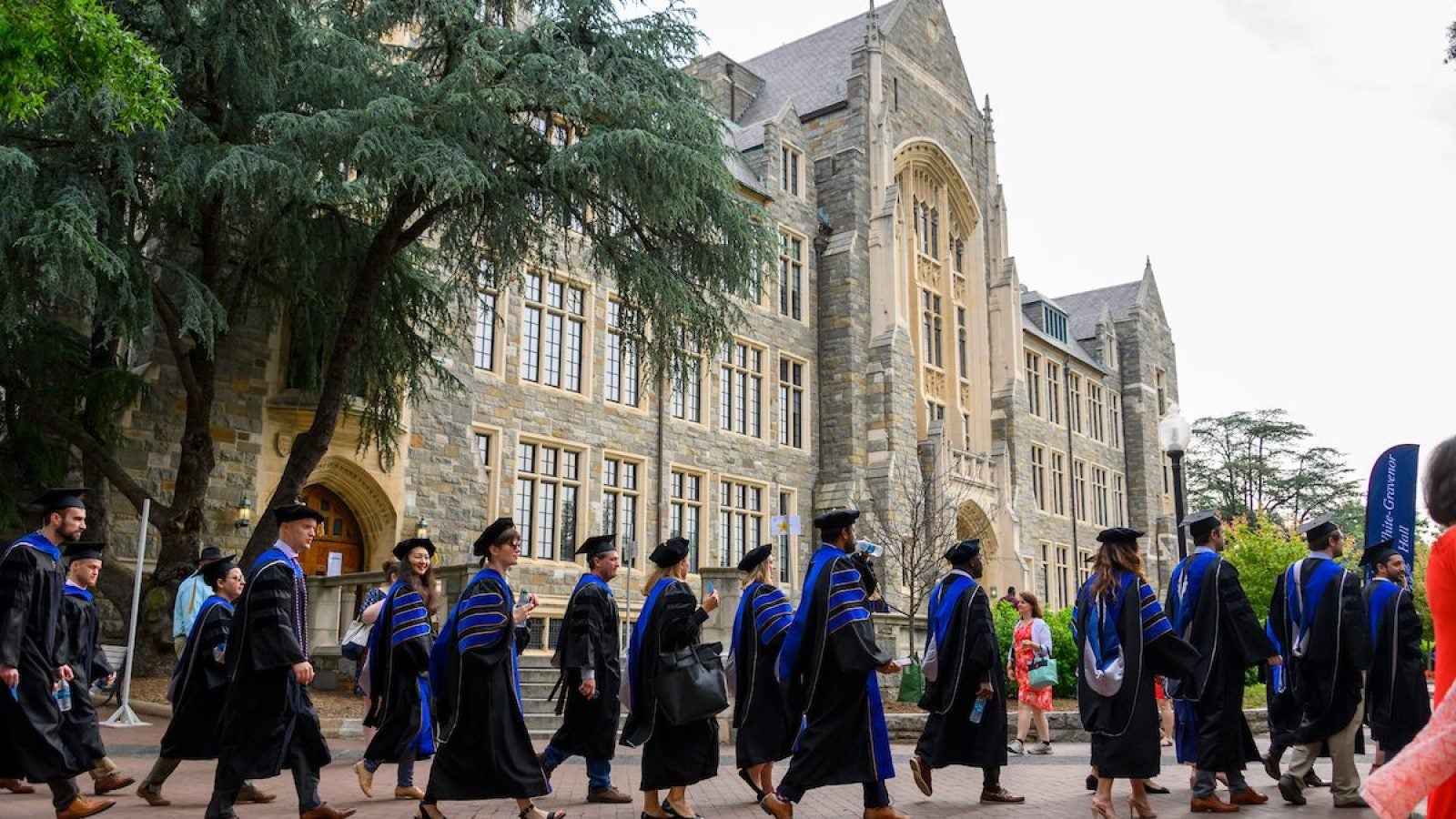 Doctoral students wearing formal black graduation gown with blue stripes on sleeves wait in line outside with White-Gravenor Hall in the background