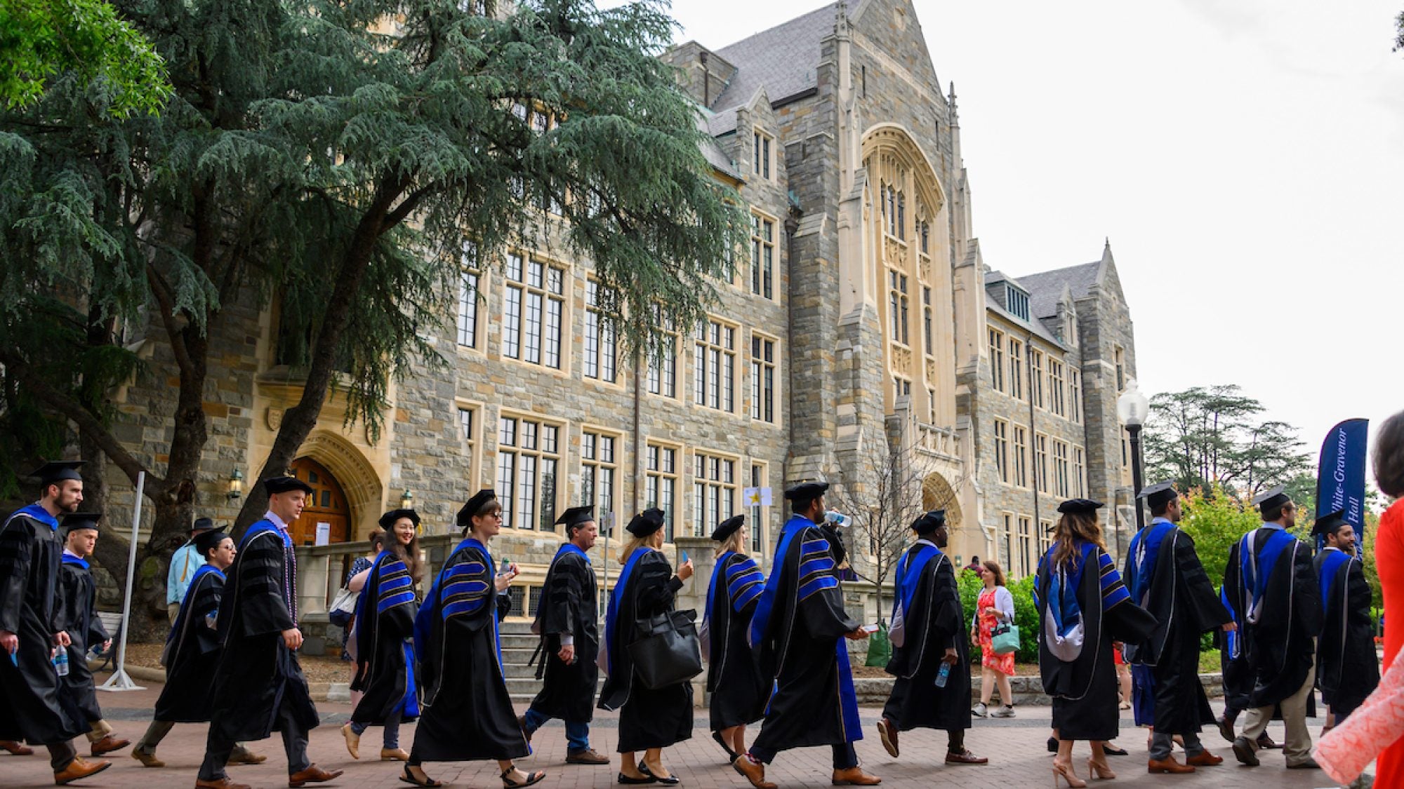 Doctoral students wearing formal black graduation gown with blue stripes on sleeves wait in line outside with White-Gravenor Hall in the background