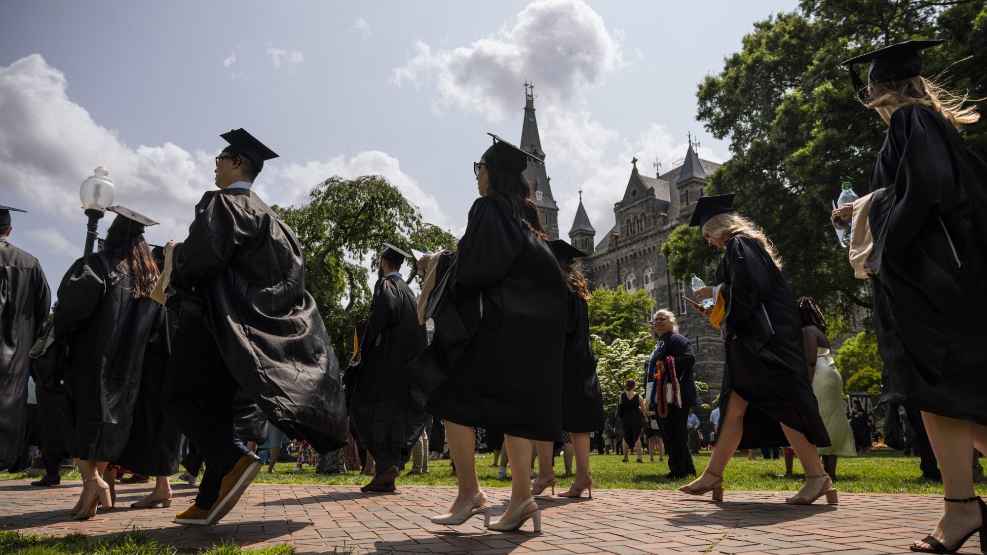 Students in black graduation robes stroll on brick walkway with Healy Hall in the background at Georgetown University