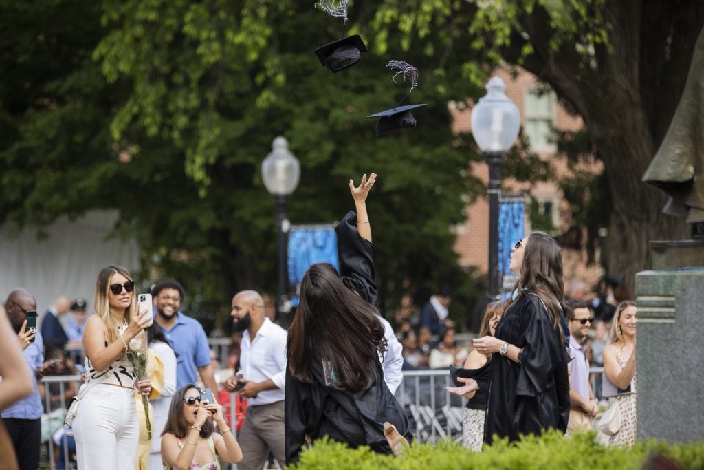 Two students in black graduation gowns toss their hats into the air while others take photos at Georgetown University