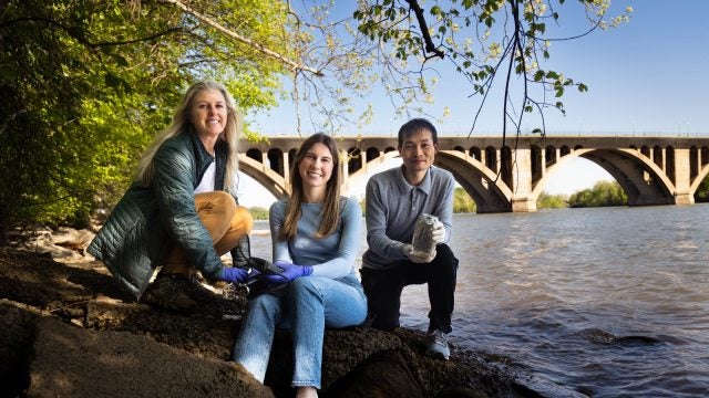 Two faculty and a student sit on the bank of Roosevelt Island in Washington, DC, with Key Bridge and Potomac River in the background