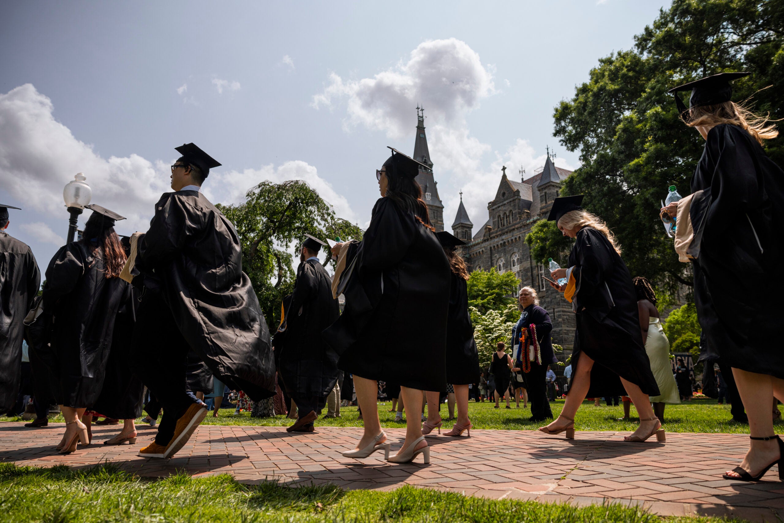 Students in black graduation robes stroll on brick walkway with Healy Hall in the background at Georgetown University