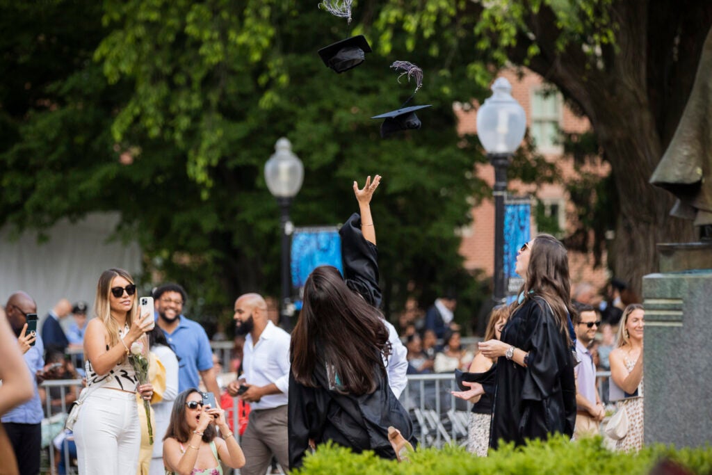 Two students in black graduation gowns toss their hats into the air while others take photos at Georgetown University