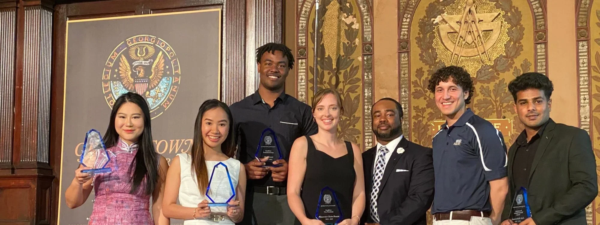 Six graduate students and a staff member pose with glass awards in Gaston Hall at Georgetown University