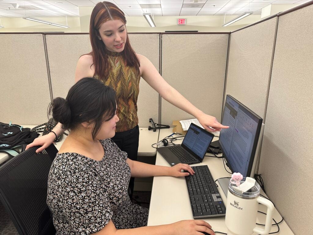 Hannah Kim (seated) sits in front of a computer with her supervisor (standing) pointing at the desktop screen