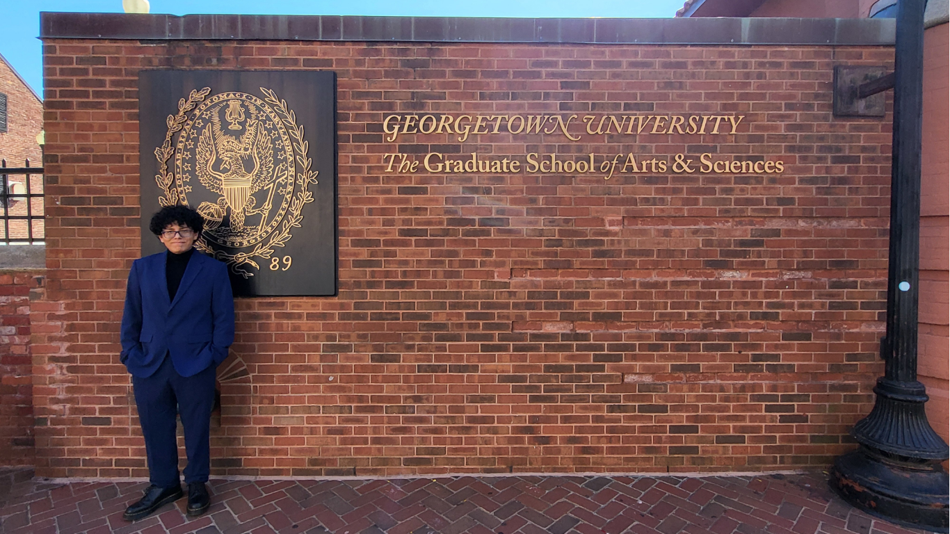 Patrick Grey (left) stands in front of the Car Barn building at Georgetown with the Graduate School seal on a brick wall; he is wearing a blue suit