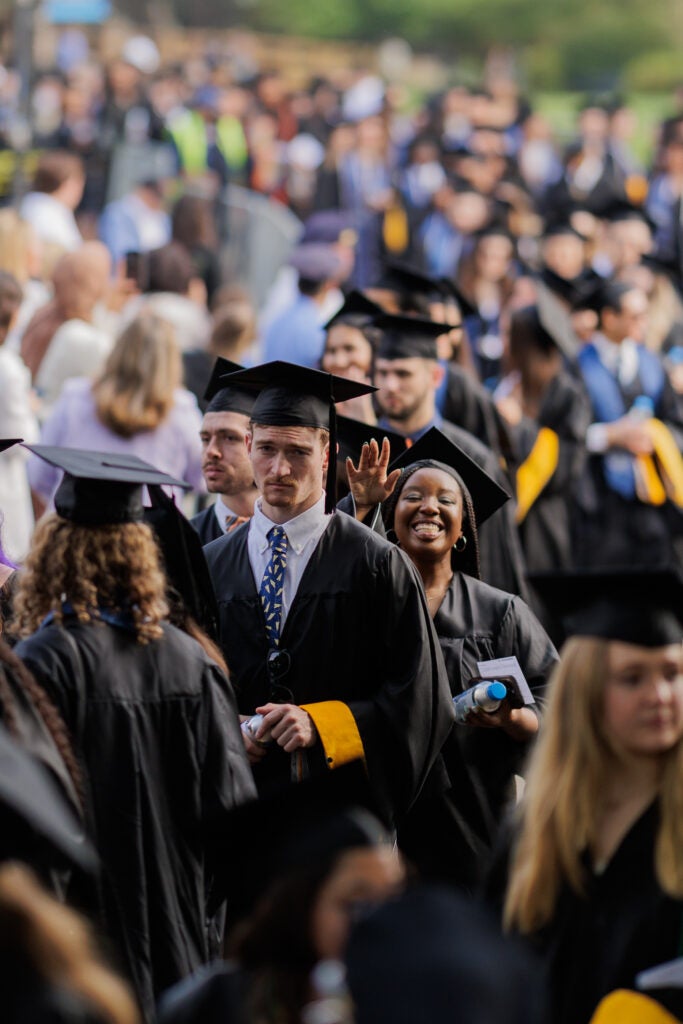Students, some waving toward the camera, line up to take their seats at Commencement 2025.