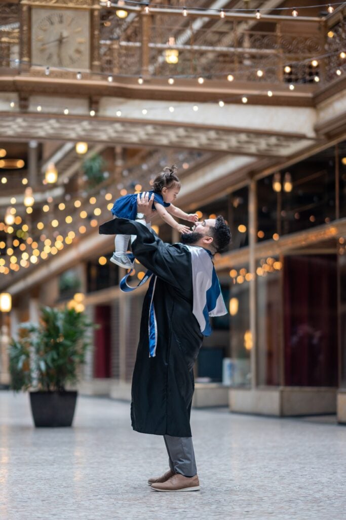 Jauregui, wearing graduation regalia, holds toddler-aged Soledad in the air above him while smiling at her.