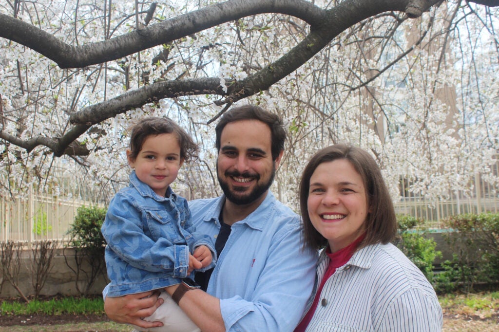 Basto, center, smiles at the camera holding his toddler daughter, Camila, to his left and with his wife, Renata, standing at his right. The family is posing in front of a blooming cherry blossom tree.