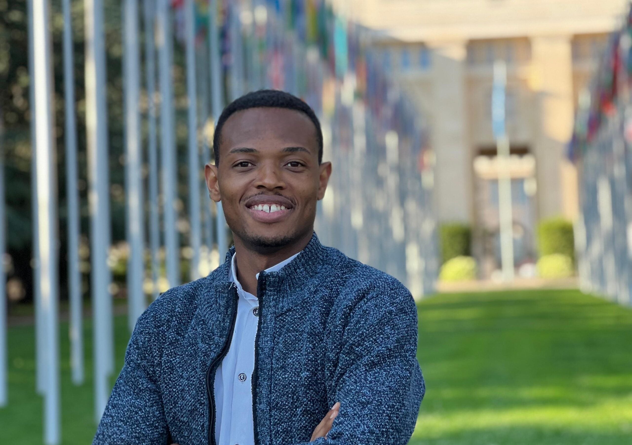 Georgetown computer science Ph.D. student Wisdom Obinna stands on a lawn in front of the U.N. office in Geneva. Behind him are flag poles flying various countries' flags.