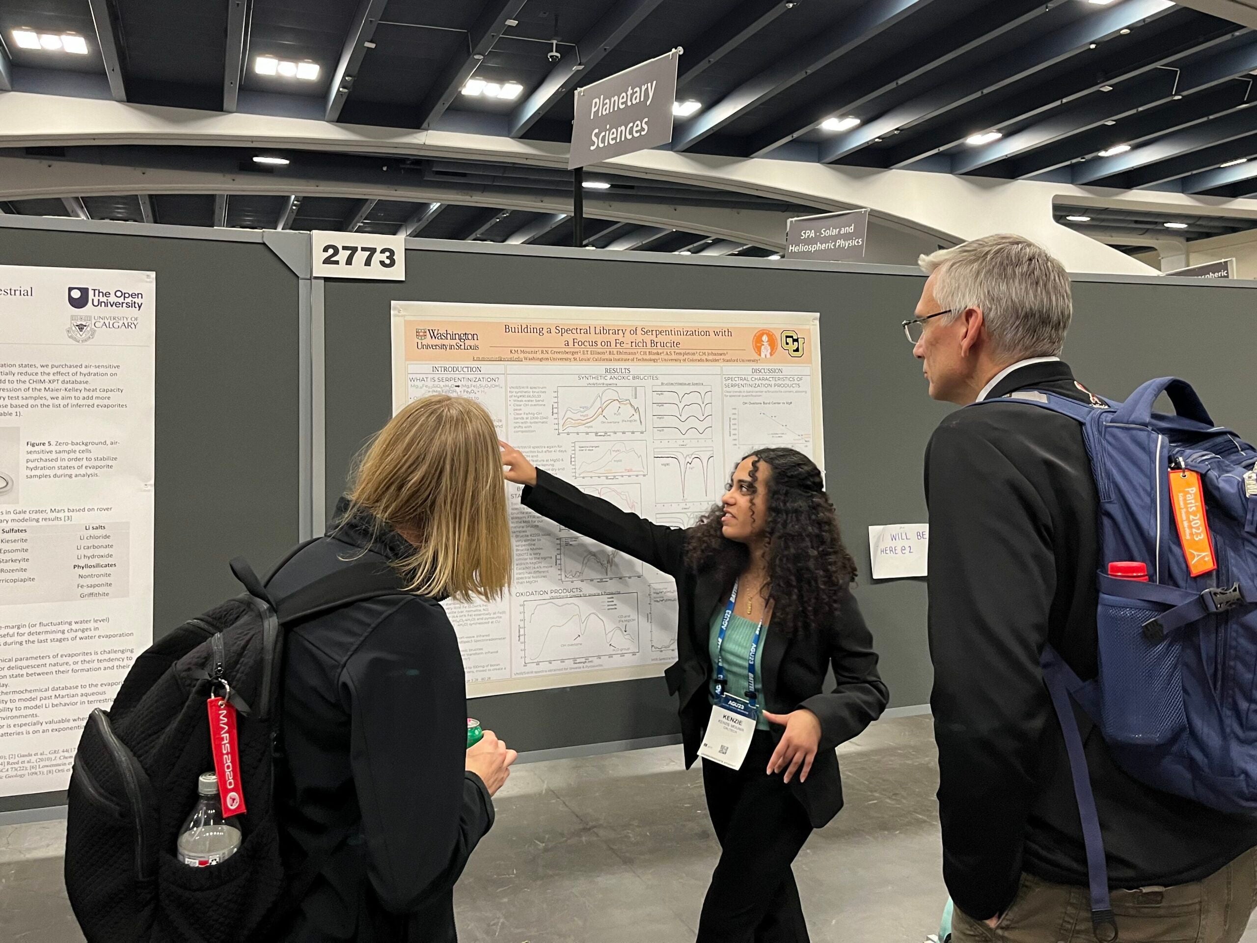 Kenzie Mounir points at a research presentation poster while speaking with two conference attendees.