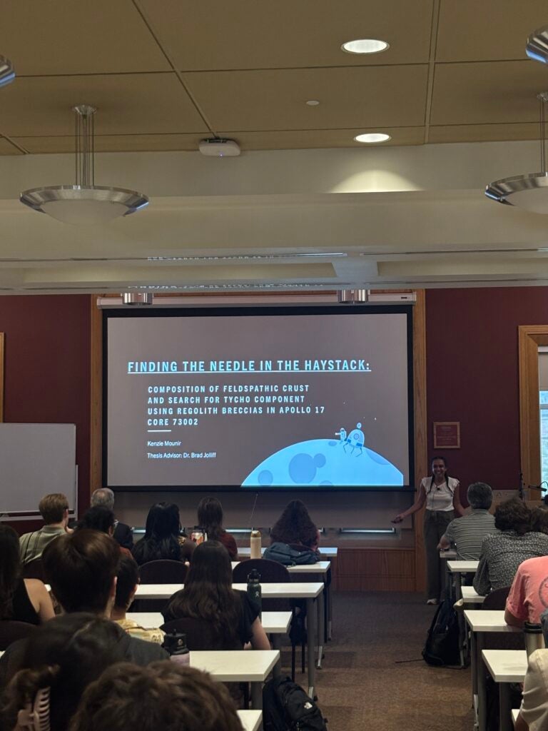 Mounir stands at the front of a classroom, to the right of a projected screen reading: "Finding the needle in the haystack: Composition of feldspathic crust and search for Tycho component using regolith breccias in Apollo 17 core 73002." People fill several rows of desks in the photo's foreground.