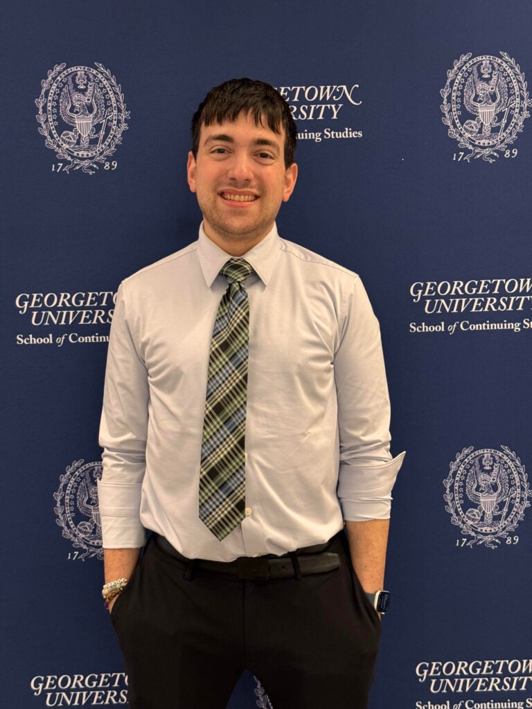 A photo of Georgetown graduate student Farrell Moskow in a dress shirt and tie in front of a photo backdrop with the Georgetown and Georgetown SCS logos.