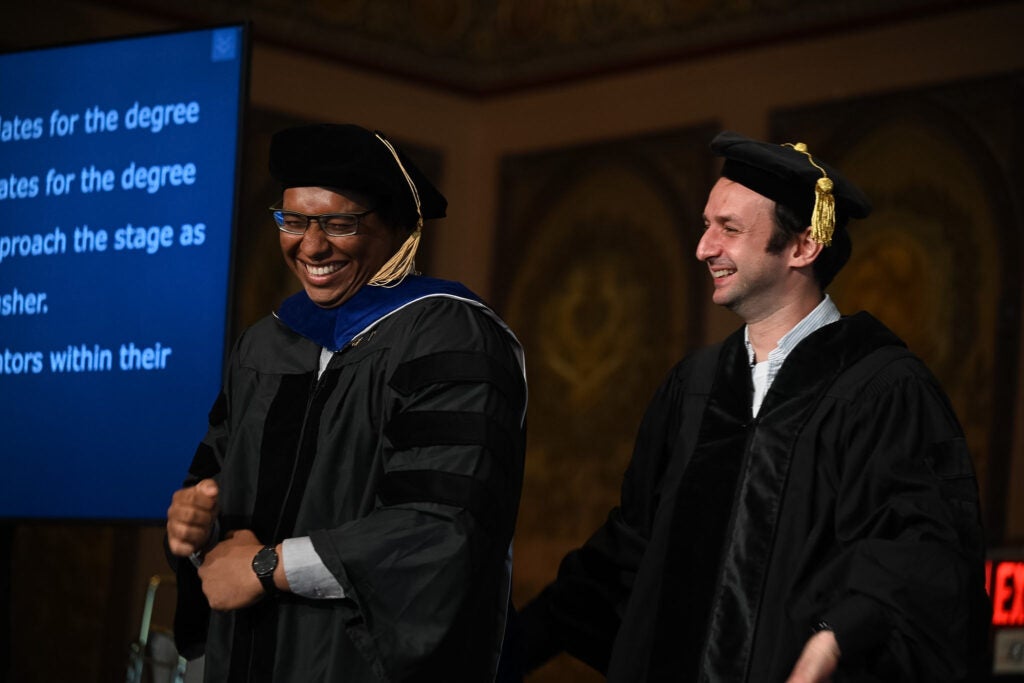 A male Ph.D. graduate grins after being hooded as his mentor looks on.