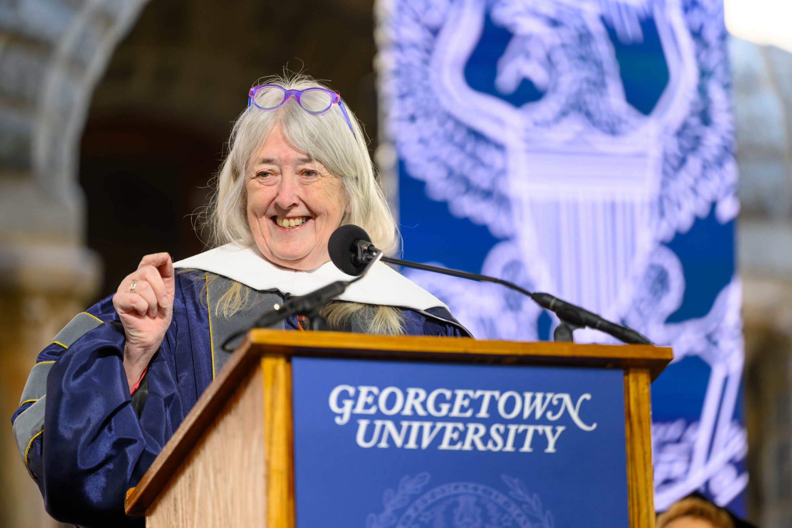 Classicist Mary Beard smiles and gestures behind a lectern with Georgetown University's logo and seal.