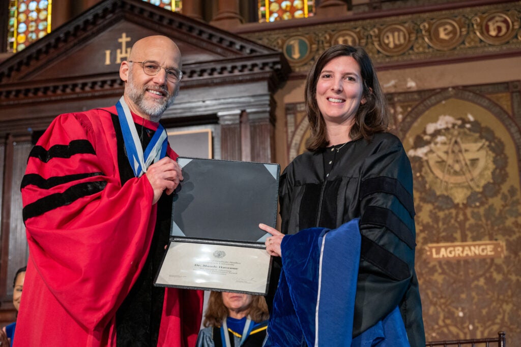 Maude Havenne, in doctoral regalia, holds her Glassman Award with Interim VP for Graduate Studies Alex Sens, also in doctoral regalia.