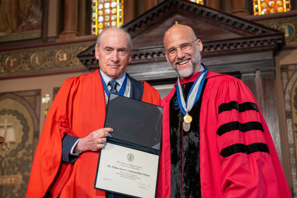 Distinguished University Professor John McNeill, dressed in doctoral regalia, holds de Vasconcellos Otoya's Glassman Distinguished Dissertation Award next to Alex Sens, vice president for Graduate Studies.