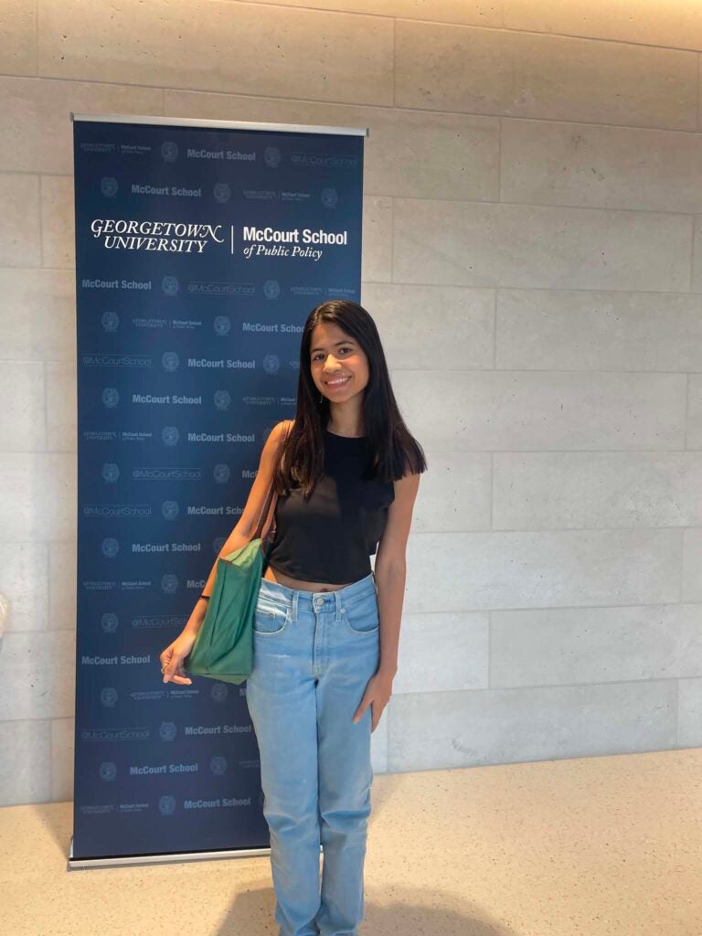 Priyasha Chakravarti poses in front of a banner reading "Georgetown University McCourt School of Public Policy"