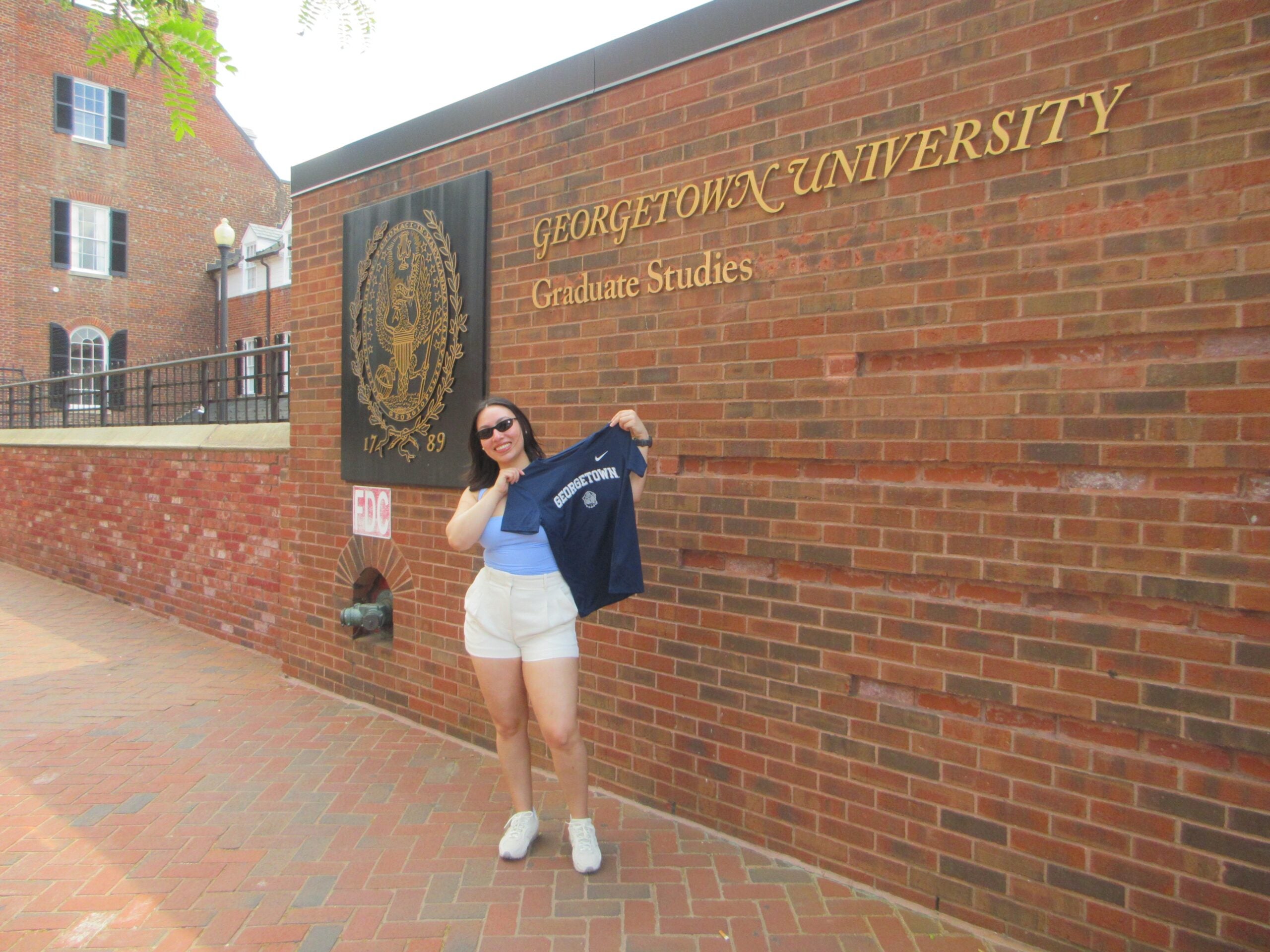 Master's student Keiti Lazo holds up a Georgetown t-shirt in front of the Graduate Studies sign on campus.