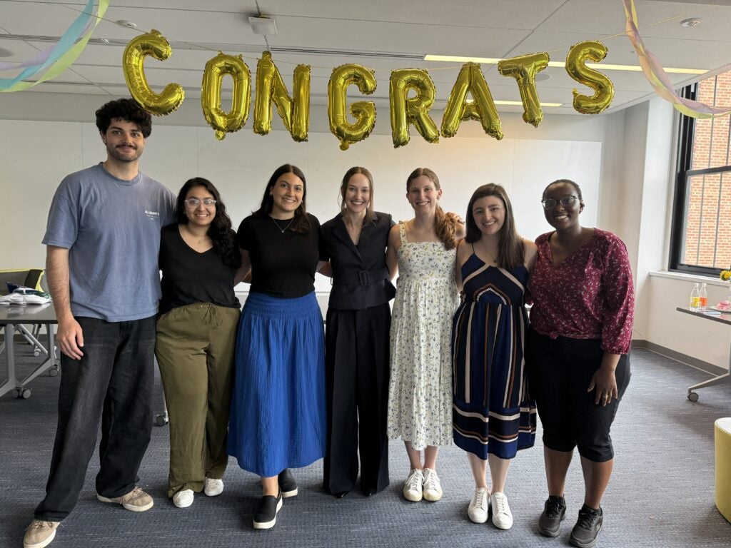 A group photo of seven people in front of a gold balloon banner reading "Congrats"