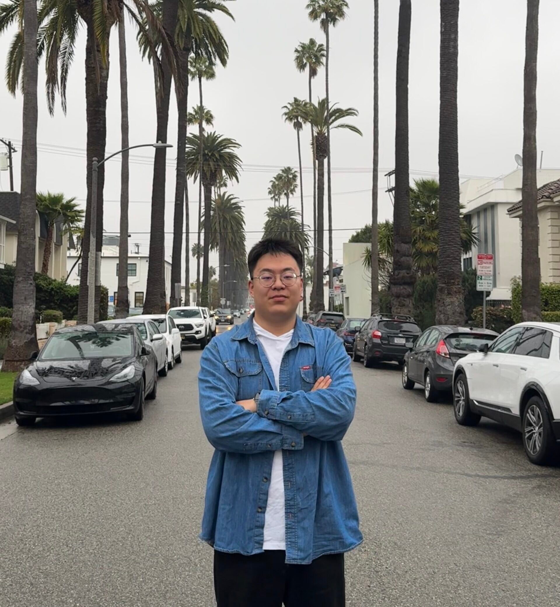 Ph.D. in biostatistics student Kaiyan Shi stands with his arms folded in front of a street background. The street is lined with palm trees.