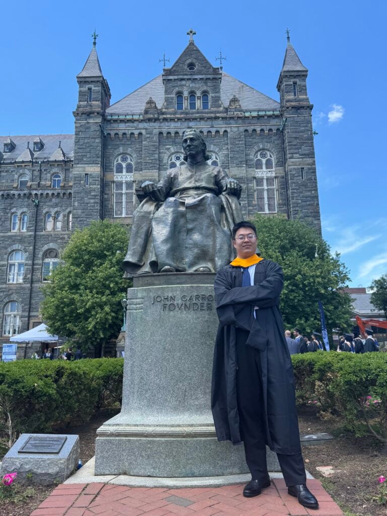 Ph.D. in biostatistics student Kaiyan Shi stands in front of the John Carroll statue near Healy Hall. Shi is wearing graduation regalia.