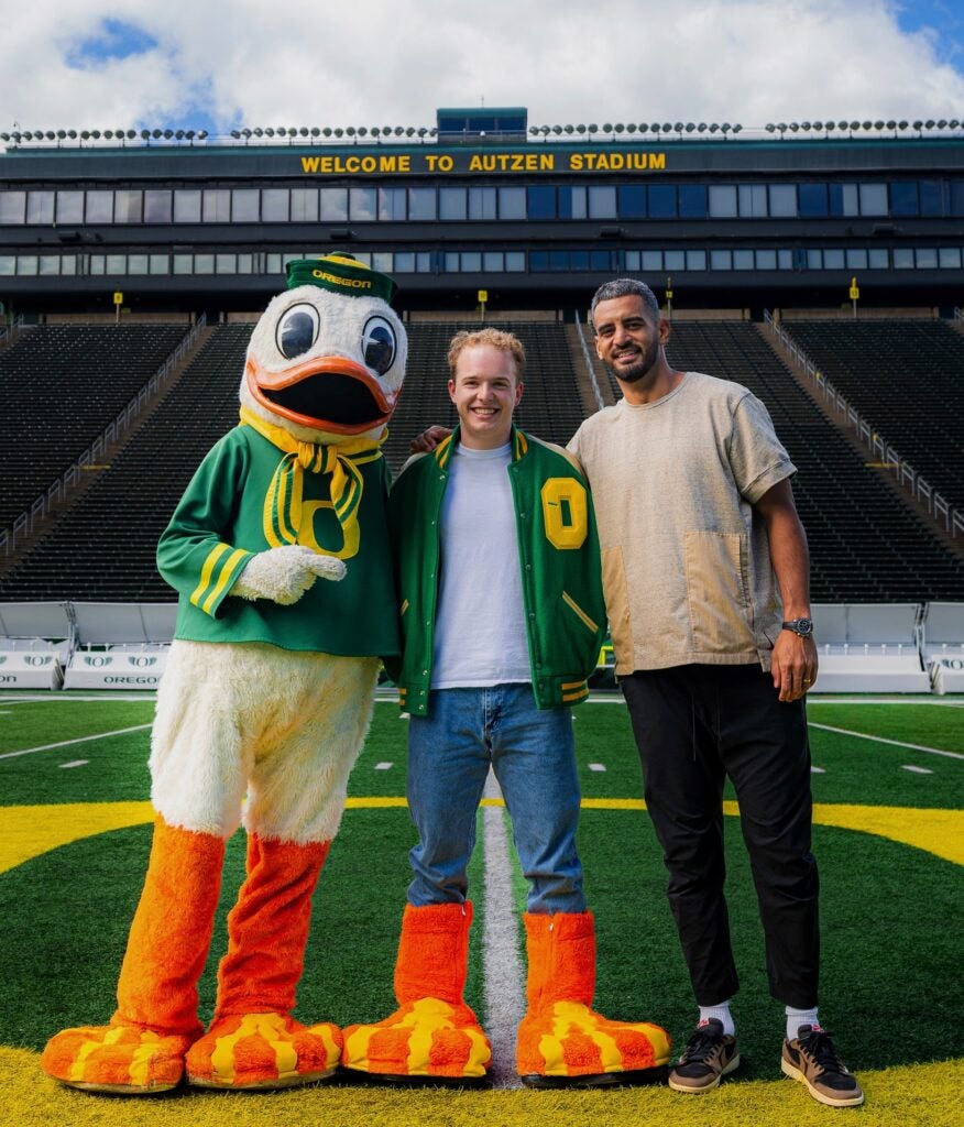 Inside the Oregon football stadium, Henry Schadwinkel wears the webbed boots of the Oregon Duck, with the Oregon Duck to his right and Washington Commanders quarterback Marcus Mariota to his left.