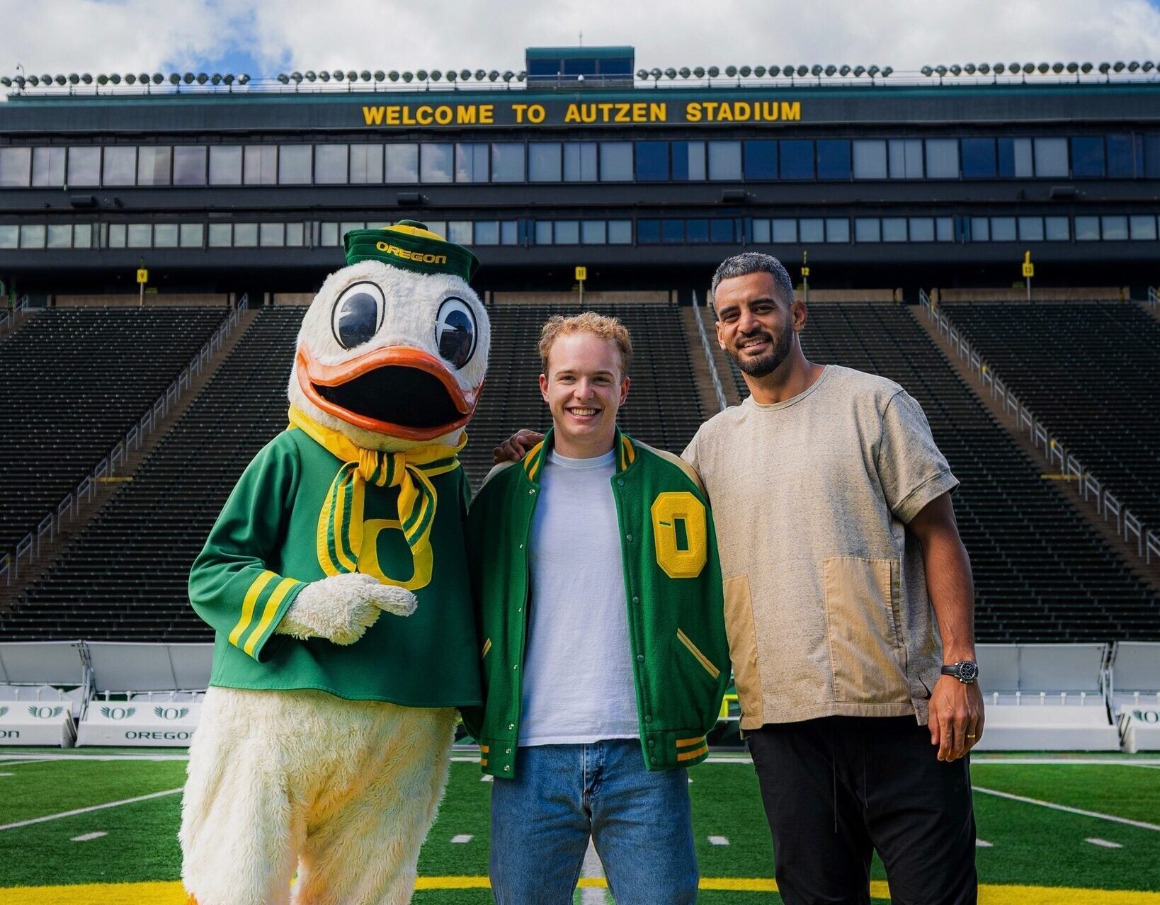 Inside the Oregon football stadium, Henry Schadwinkel wears the webbed boots of the Oregon Duck, with the Oregon Duck to his right and Washington Commanders quarterback Marcus Mariota to his left.