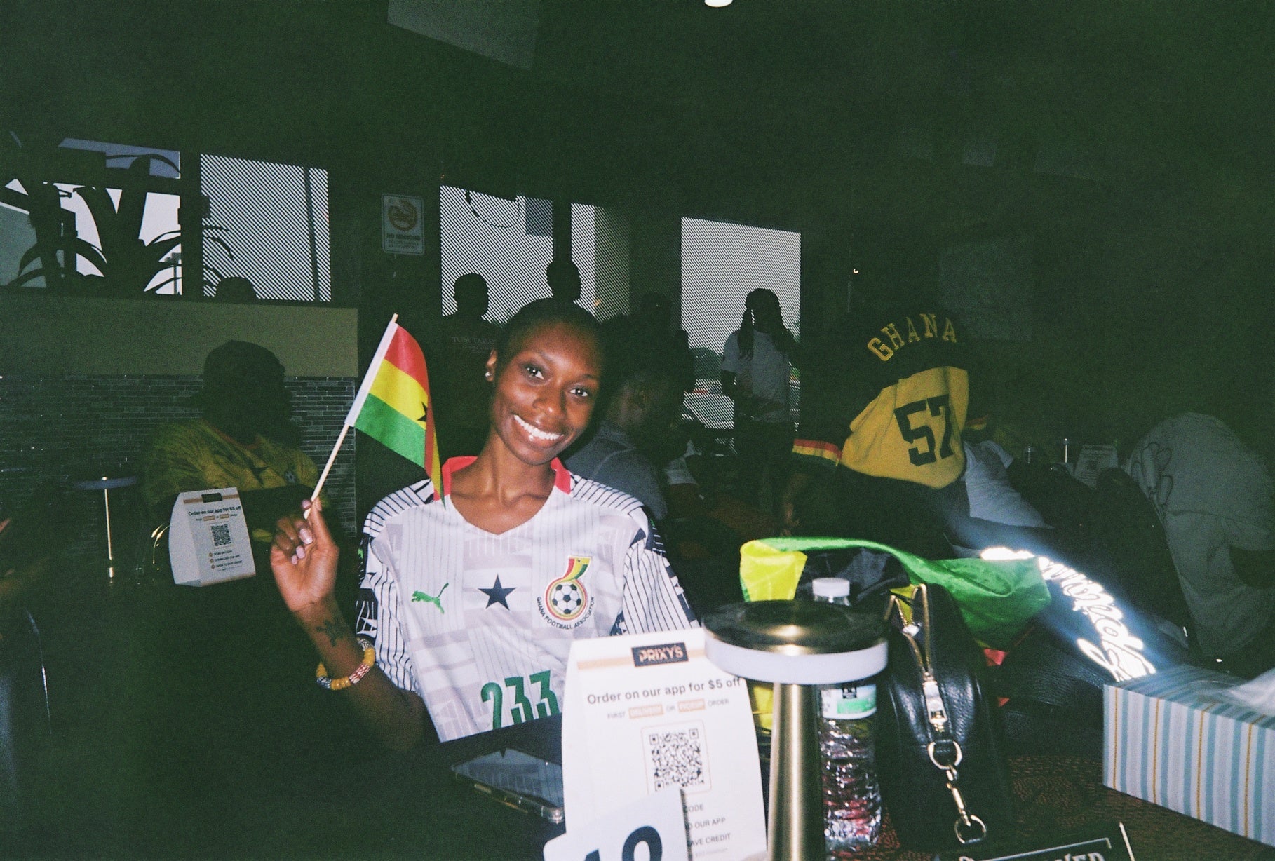 Jennifer Ayarkwa holds a red-, yellow- and green-striped flag while wearing a sports jersey and sitting at a table in a busy restaurant.