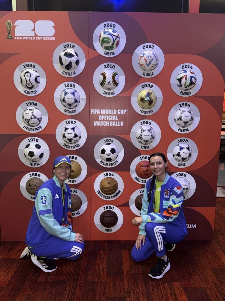 Georgetown student Rachel Beatty, right, kneels in front of a display of FIFA World Cup Official Match Balls. Another volunteer poses to her right.